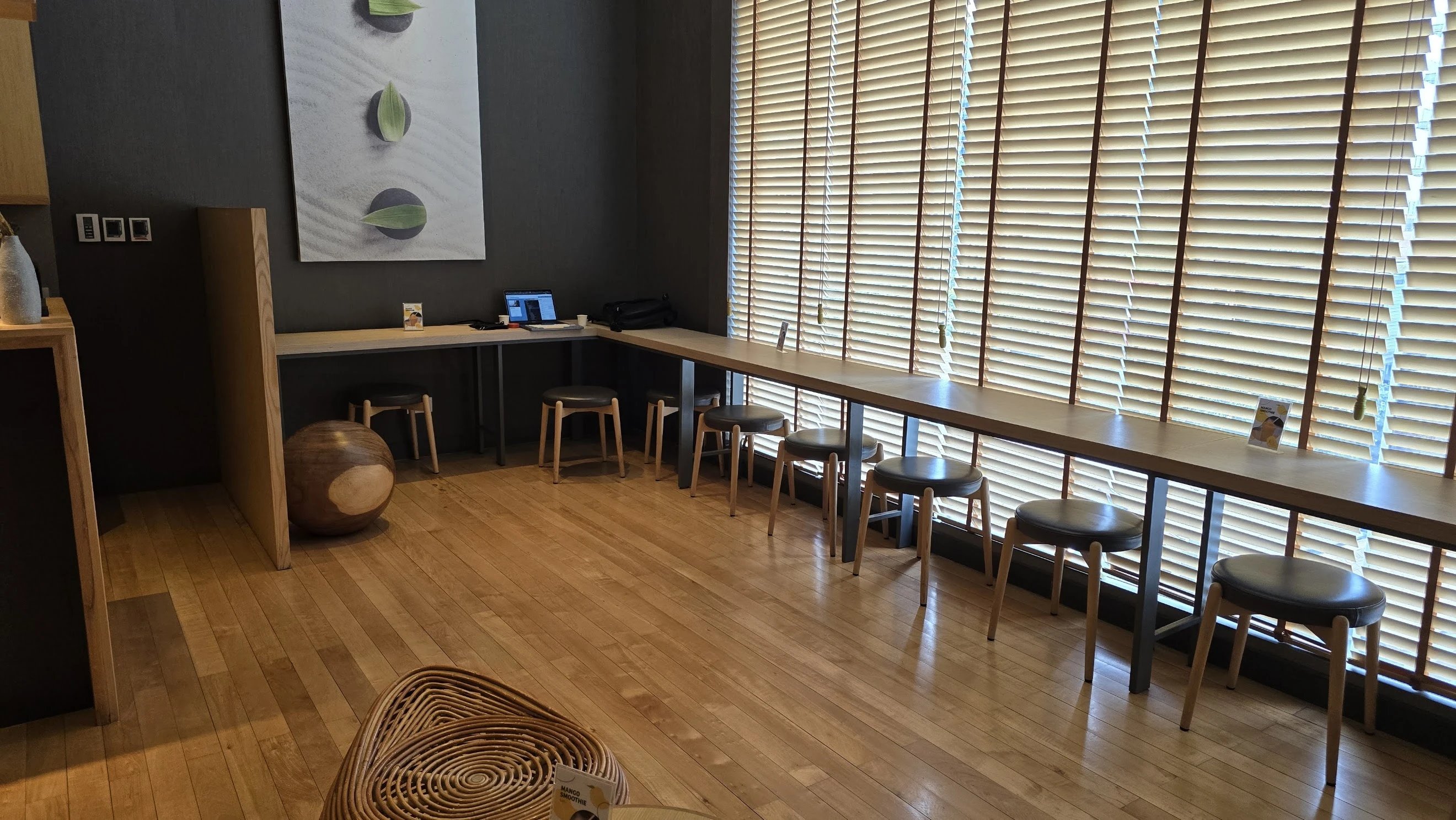 A modern and minimalist cafe interior with wooden floors, a row of stools along a counter, and large windows with blinds providing natural lighting.