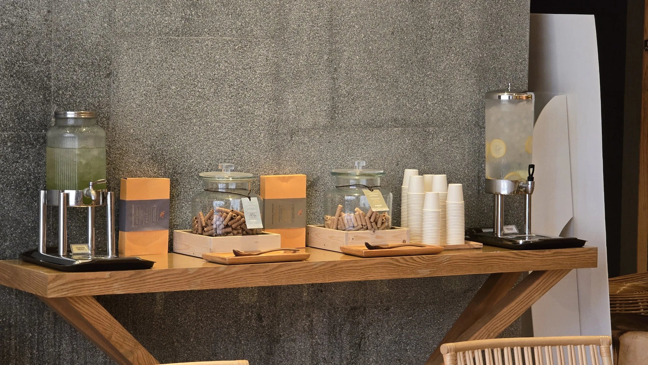 A wooden shelf displays various items, including containers for pandan water and infused water, coconut rolls, and paper cups, set against a gray concrete wall background.